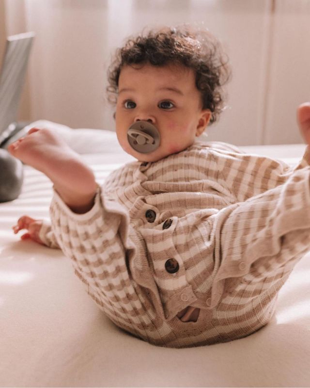 BIBS COLOUR Bebé com fato às riscas bege e chupeta cinzenta deitado numa cama branca
