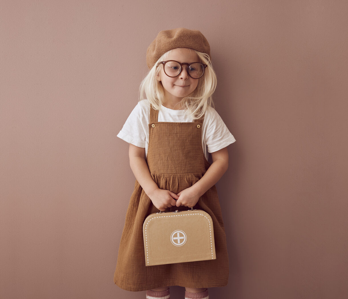 KIT MÉDICO Menina loira com óculos, boina castanha e vestido castanho segurando mala pequena