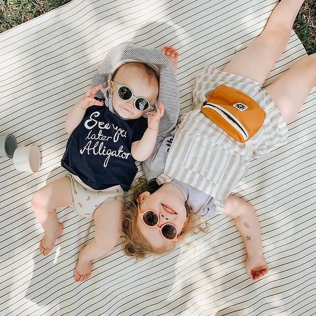 ÓCULOS IZIPIZI | KIDS Duas crianças deitadas numa manta riscada com roupa colorida e óculos de sol