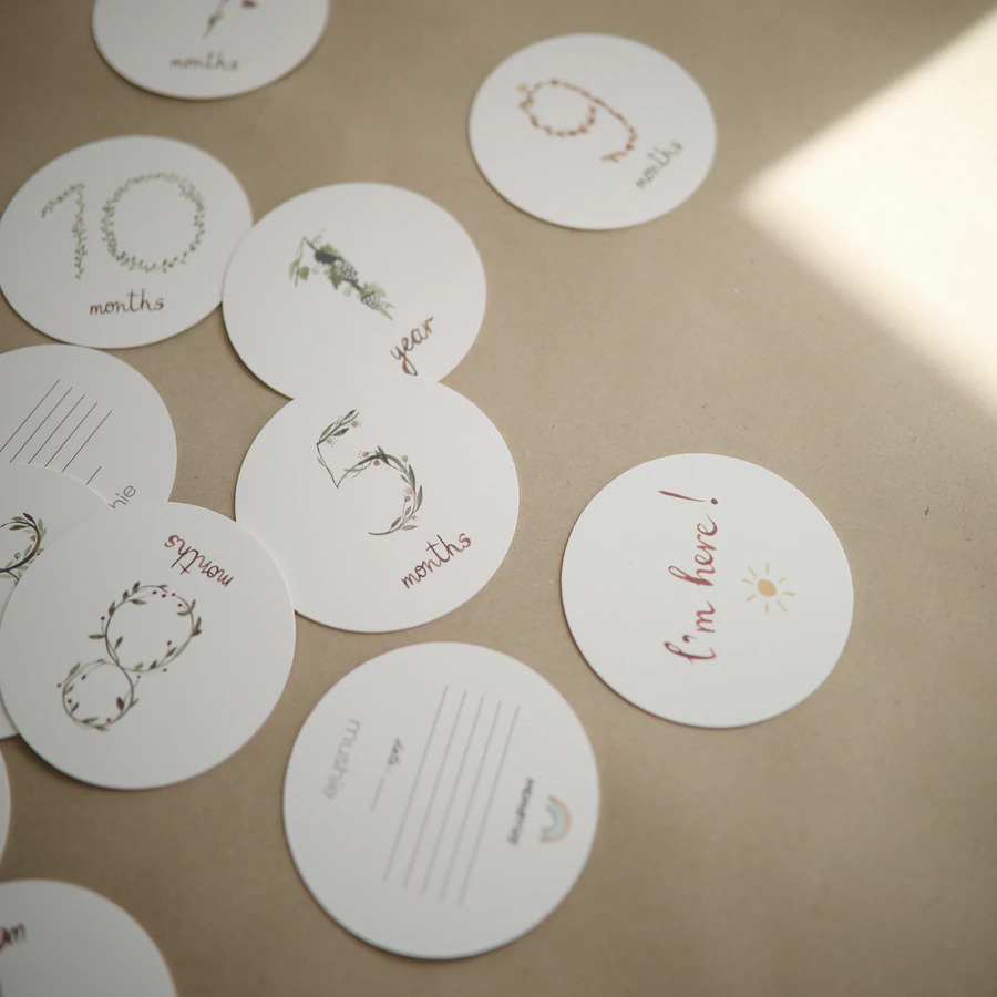 CARTÕES MILESTONE Cartões redondos brancos com números e textos indicando meses e idades sobre uma superfície bege