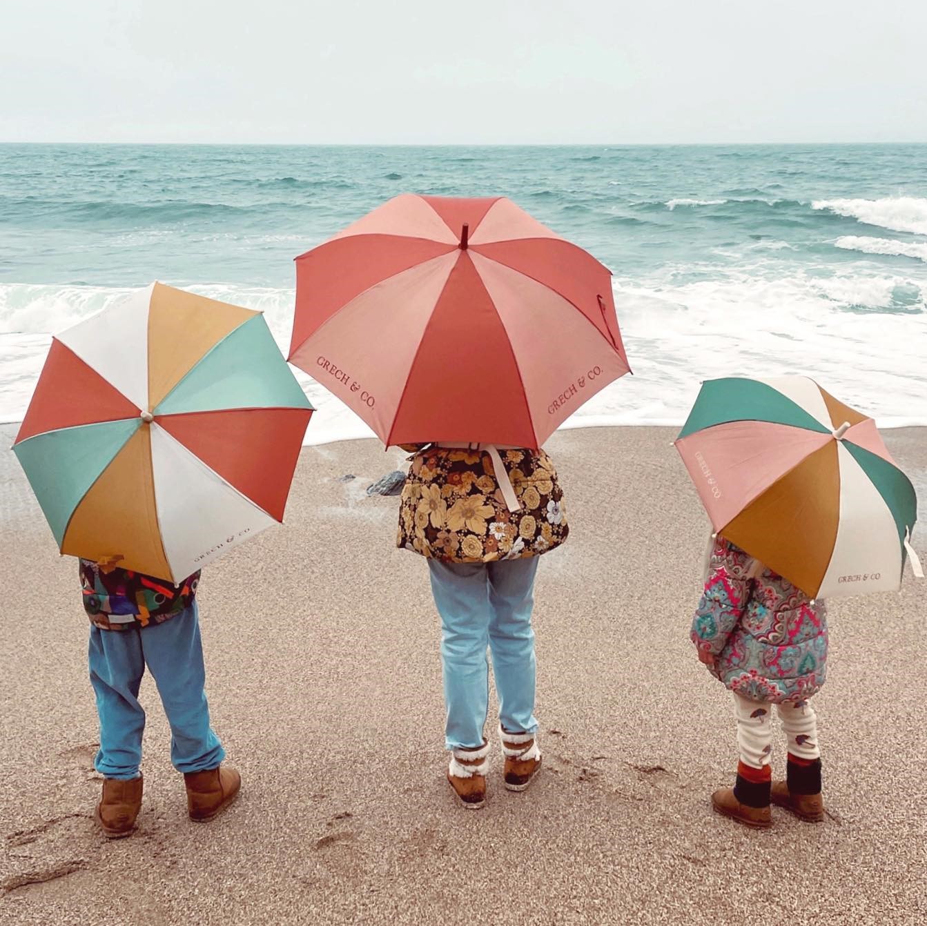 CHAPÉU DE CHUVA SUSTENTÁVEL | MOMMY & ME Três crianças com guarda-chuvas coloridos na praia