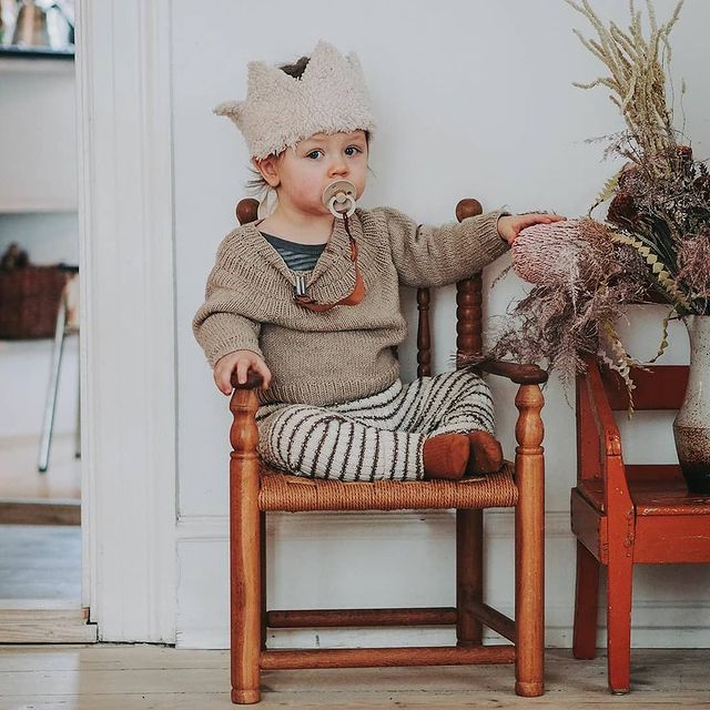 BIBS COLOUR Criança sentada numa cadeira de madeira com roupa de malha e chapéu em forma de coroa