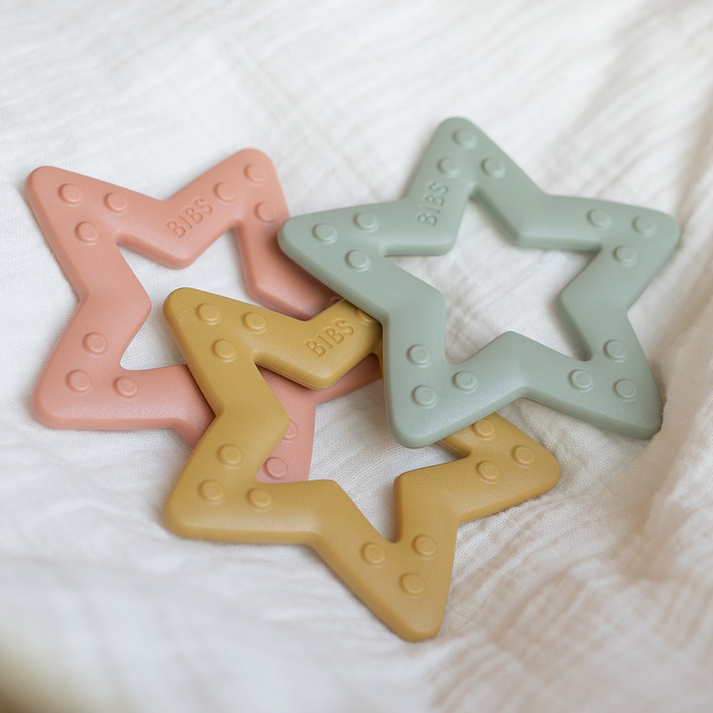 MORDEDORES | BIBS Quatro mordedores em forma de estrela de silicone em cores pastel colocados sobre tecido branco.