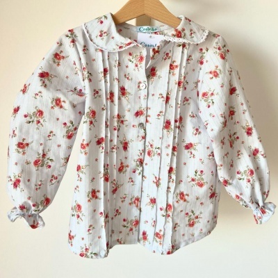 Camisa infantil branca com flores vermelhas e rosas, mangas compridas, gola redonda, pendurada num cabide de madeira