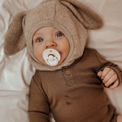 Bebé com chapéu felpudo com orelhas e fato de malha castanho deitado em cama branca