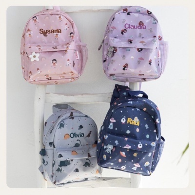 Quatro mochilas infantis coloridas com nomes bordados penduradas numa escada branca