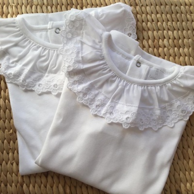 Dois bodies brancos de bebé com gola de renda sobre superfície de juta trançada