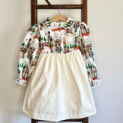 Vestido infantil com estampado de soldadinhos de chumbo e inverno em fundo branco, saia e gola brancas, pendurado em estrutura de madeira.