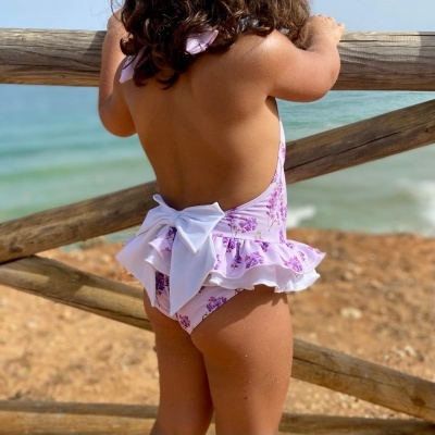 Menina de costas com fato de banho lilás claro e detalhes em branco, encostada a vedação de madeira junto ao mar