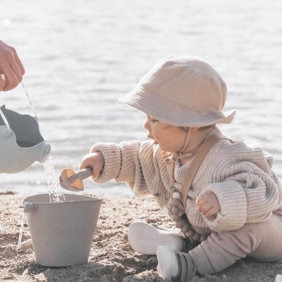 Criança a brincar na praia com balde e pá, vestida de malha e chapéu bege.