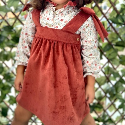 Criança usando vestido vermelho de veludo com camisa floral em fundo de cerca de madeira e vegetação