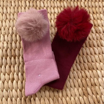 Duas meias com pompom em malha, uma rosa e outra vinho, sobre superfície trançada