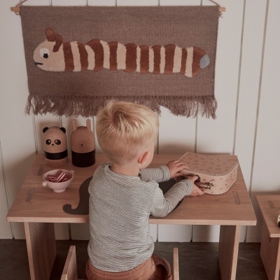 Criança sentada numa secretária de madeira com caixa estampada e tapeçaria de animal listrado na parede.