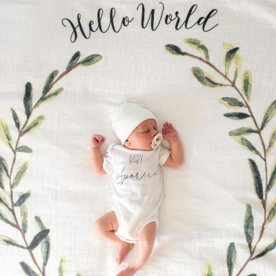 Bebé com body branco e gorro deitado numa manta branca com texto e folhas verdes.