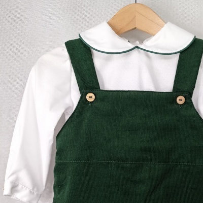 Roupa de criança com camisa branca e jardineira verde escura pendurada num cabide.