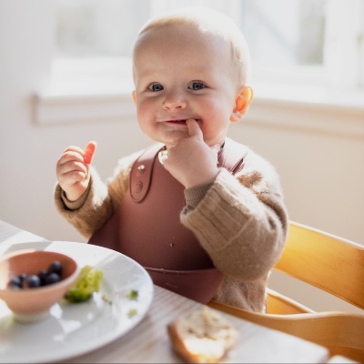 Bebé com babete de silicone castanho sentado à mesa com comida