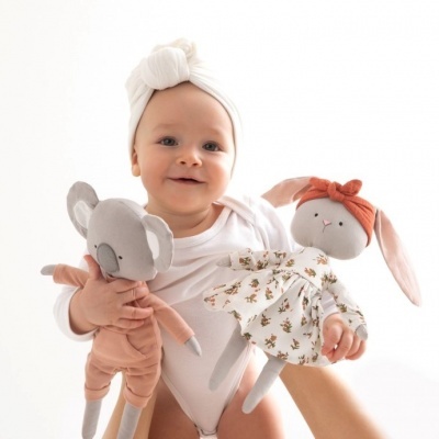 Bebé com body branco e touca branca a segurar duas bonecas de peluche, coala e coelha.