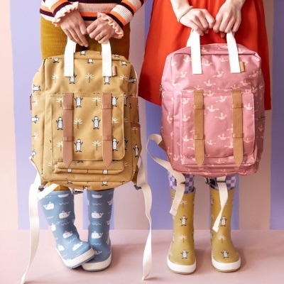 Duas mochilas retangulares coloridas com padrões infantis seguradas por crianças com botas de chuva coloridas em fundo pastel.