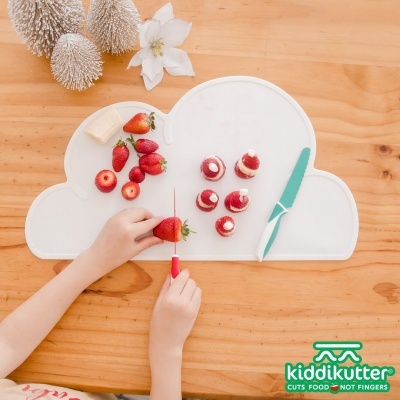 Tapete de silicone branco em forma de nuvem com morangos cortados e uma faca verde numa mesa de madeira