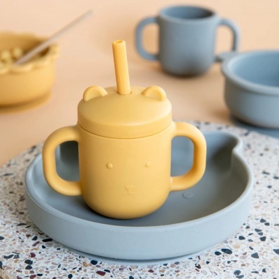 Copo de silicone amarelo com tampa e palhinha em prato azul claro sobre mesa de superfície terrazzo