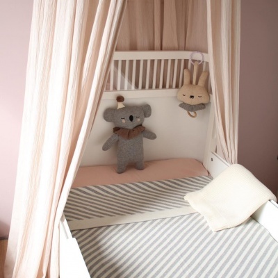 Berço branco com cortinas cor de rosa, colchão rosa, cobertor cinza riscado, almofada branca e brinquedos macios