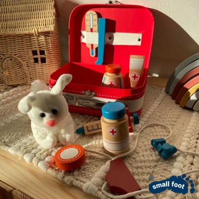 Conjunto de brinquedo médico com mala vermelha, acessórios e coelho de pelúcia sobre tapete bege