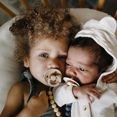 Duas crianças pequenas com chupetas, uma com roupa branca e a outra com colar de contas, sentadas numa almofada branca