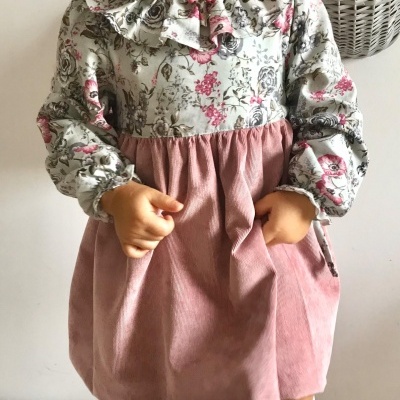 Bebé com vestido floral e cor-de-rosa, interior com cesta cinzenta ao fundo.