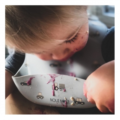 Criança com babete de silicone branco com carrinhos e tratores, com manchas de comida roxa.