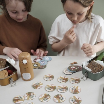 Crianças a jogar com cartas redondas ilustradas, com lancheiras verdes e garrafa térmica bege em forma de urso