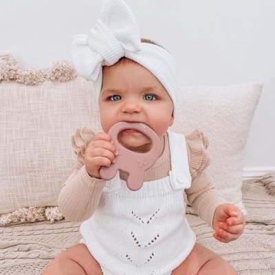 Bebé com roupa branca e faixa com laço a morder chupeta cor de rosa sentado em manta bege.