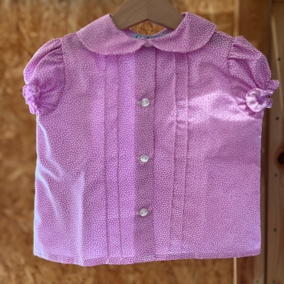 Blusa infantil rosa com pequenos pontos brancos e botões brancos, pendurada num cabide de madeira