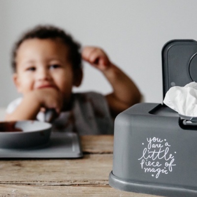 Dispensador cinzento para toalhetes para bebé com texto branco, numa mesa de madeira com bebé ao fundo
