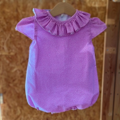 Macacão de bebé rosa com gola folhada e padrão de pontos brancos pendurado em cabide de madeira