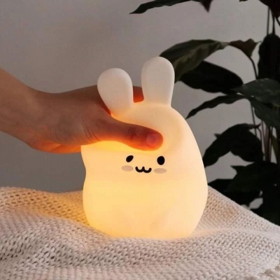 Luminária de coelho macio com luz amarela em tecido branco, com planta ao fundo