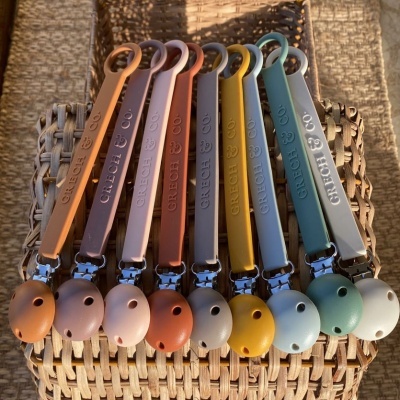 Oito presilhas para chupetas em várias cores organizadas sobre superfície de vime e fundo de pedra