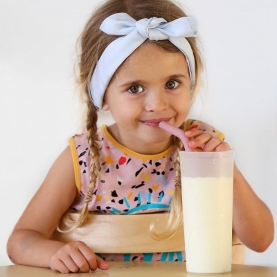 Criança loira com fita branca a beber leite num copo alto com canudo rosa.