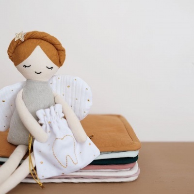 Boneca anjo de pano segurando bolsa branca com padrão de dente dourado sobre tecidos coloridos