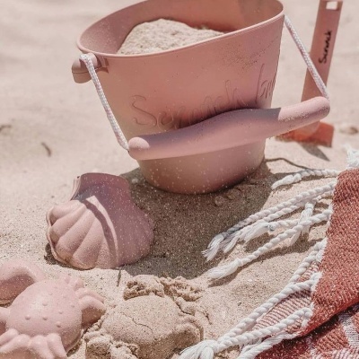 Brinquedos de praia cor-de-rosa em areia junto a toalha com franjas
