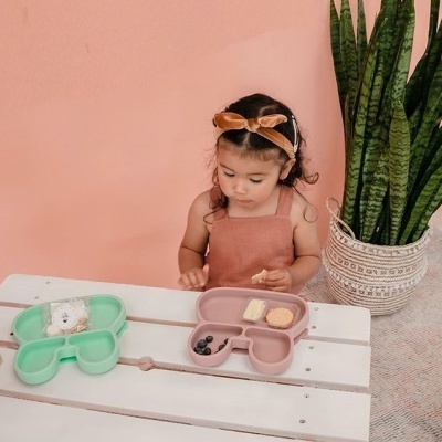 Criança com pratos para snacks em forma de urso numa mesa branca, parede rosa e planta verde