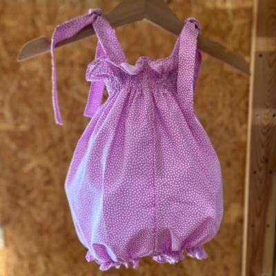 Macacão curto lilás com pontos brancos para bebé pendurado em cabide de madeira