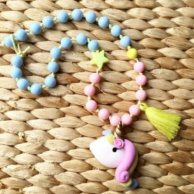 Colar colorido com pendente de unicórnio branco e rosa, e tassel amarelo, sobre superfície trançada