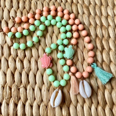 Colares coloridos com contas e pendentes de tartaruga e concha, sobre tecido trançado de fibra natural.