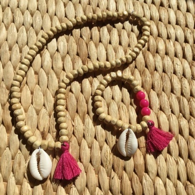 Conjunto de colar e pulseira com contas bege, conchas brancas e pompons rosa escuro sobre superfície de palha trançada.