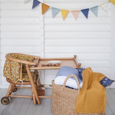 Cadeira de criança em madeira clara com almofada, cesta de palha com almofadas e manta e bandeirolas coloridas na parede