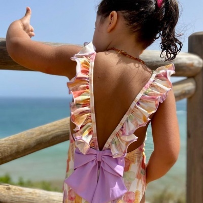 Vestido infantil com padrão floral, folhos e laço lilás nas costas, vista de costas com mar ao fundo