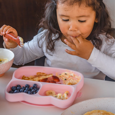 Prato infantil cor-de-rosa subdividido com panquecas e frutas, criança a comer numa mesa branca