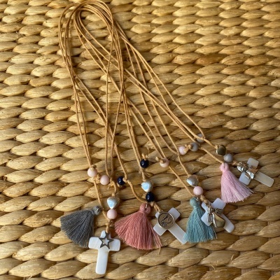 Colares com cruz branca, contas coloridas e tasséis em tons pastéis sobre superfície de fibra natural