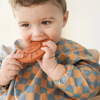 Criança a morder brinquedo de silicone alaranjado com padrão de sol, vestindo babete xadrez azul e castanho