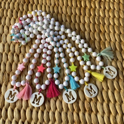 Pulseiras com contas brancas, estrelas coloridas, pompons e medalhões com letra M sobre tecido tramado.